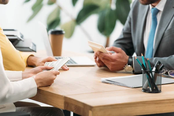 Abgeschnittene Ansicht einer Frau, die am Tisch sitzt und Dollarnoten in der Nähe eines Mannes hält, wile Berater, der Taschenrechner im Büro hält — Stockfoto