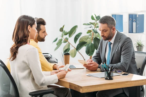 Woman sitting at table near man and holding dollar banknotes wile advisor holding calculator in office — Stock Photo