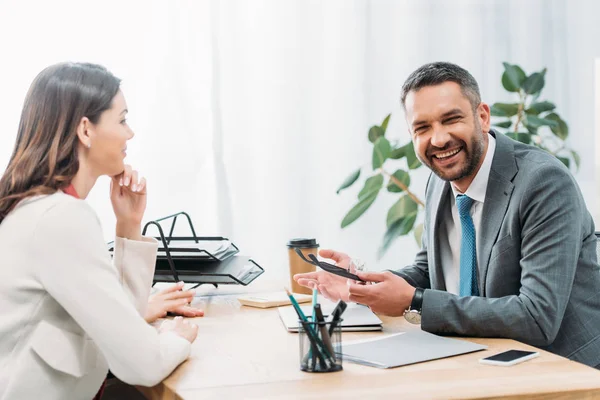 Conselheiro sentado à mesa sorrindo e segurando óculos com investidor no escritório — Fotografia de Stock