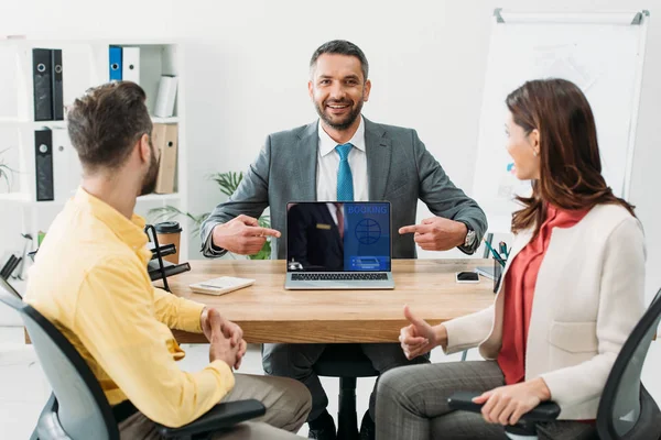 Conseiller pointant avec les doigts à l'ordinateur portable avec le site de réservation à l'écran près de l'homme et la femme pouce vers le haut dans le bureau — Photo de stock