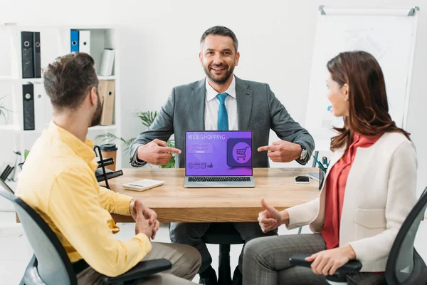 Advisor pointing with fingers at laptop with web shopping website on screen near man and woman thumbing up in office — Stock Photo