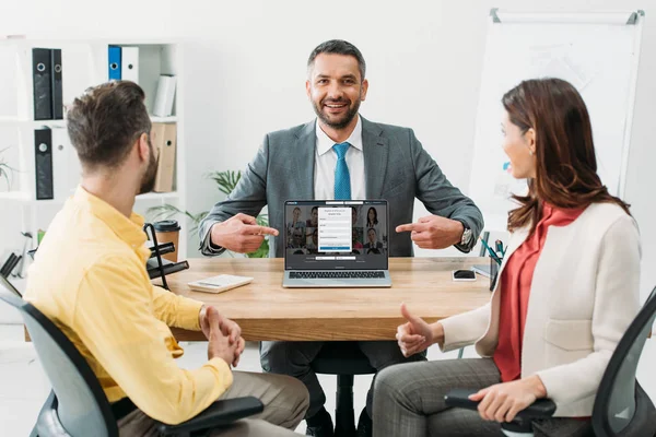 Asesor apuntando con los dedos a la computadora portátil con linkedin sitio web en la pantalla cerca de hombre y mujer pulgar hacia arriba en la oficina - foto de stock