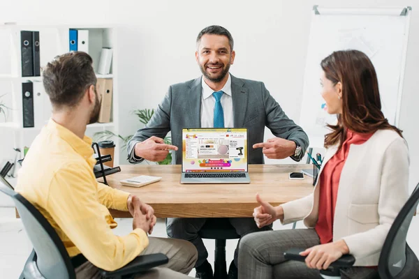 Conselheiro apontando com os dedos para laptop com site aliexpress na tela perto de homem e mulher batendo no escritório — Fotografia de Stock