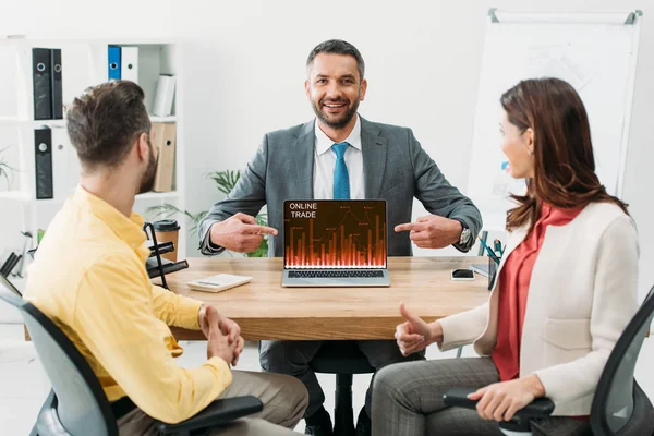 Advisor pointing with fingers at laptop with online trade website on screen near man and woman thumbing up in office — Stock Photo