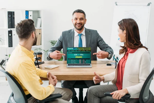 Selective focus of advisor pointing with fingers at laptop with sportsbet online website on screen to couple in office — Stock Photo