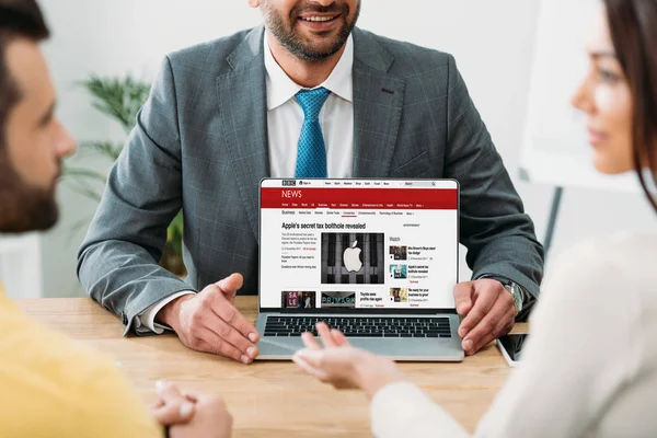 Cropped view of advisor sitting at table and laptop with bbc news website on screen with investors in office — Stock Photo