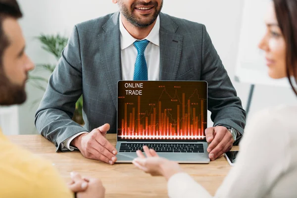 Vista recortada del asesor sentado en la mesa y el ordenador portátil con el sitio web de comercio en línea en la pantalla con los inversores en la oficina - foto de stock