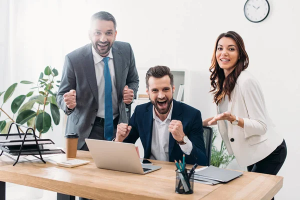 Colegas en la mesa mirando a la computadora portátil, regocijándose y sonriendo en la oficina - foto de stock