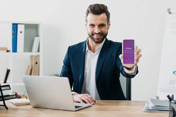 Businessman sitting at table with laptop and showing smartphone with instagram app on screen in office — Stock Photo