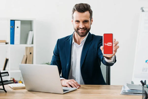 Businessman sitting at table with laptop and showing smartphone with youtube app on screen in office — Stock Photo