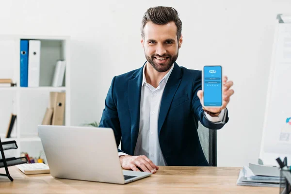 Hombre de negocios sentado en la mesa con el ordenador portátil y mostrando el teléfono inteligente con la aplicación de Skype en la pantalla en la oficina - foto de stock