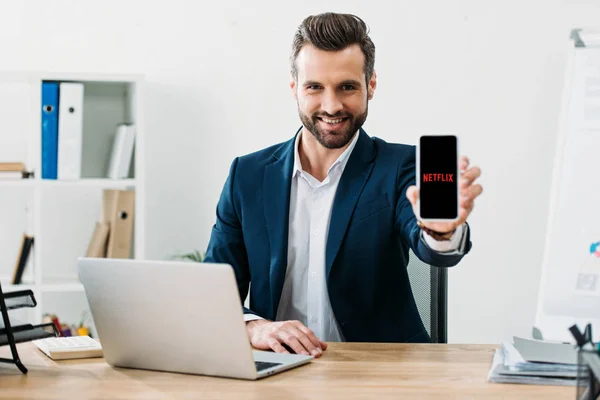 Businessman sitting at table with laptop and showing smartphone with netflix app on screen in office — Stock Photo