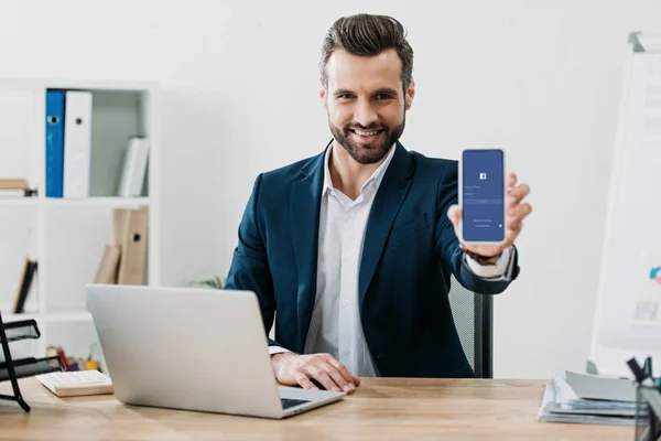 Uomo d'affari seduto a tavola con laptop e mostrando smartphone con app facebook sullo schermo in ufficio — Foto stock