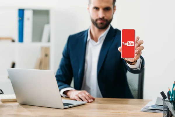 Selective focus of businessman sitting at table with laptop and showing smartphone with youtube app on screen in office — Stock Photo