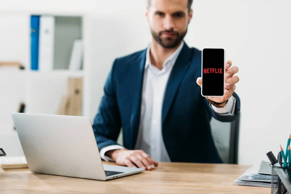 Selective focus of businessman sitting at table with laptop and showing smartphone with netflix app on screen in office — Stock Photo