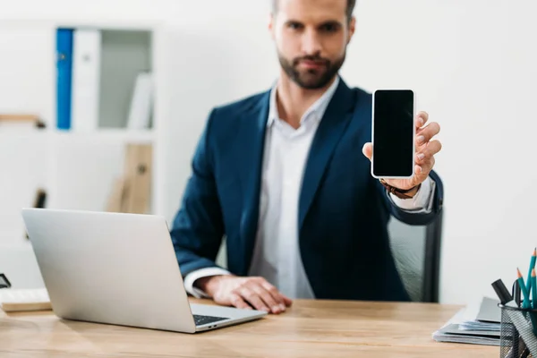Вибірковий фокус бізнесмена, що сидить за столом з ноутбуком і показує порожній екран смартфона в офісі — стокове фото