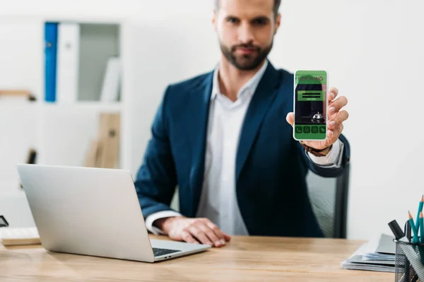 Foyer sélectif de l'homme d'affaires à table avec ordinateur portable et montrant smartphone avec application de réservation à l'écran dans le bureau — Photo de stock