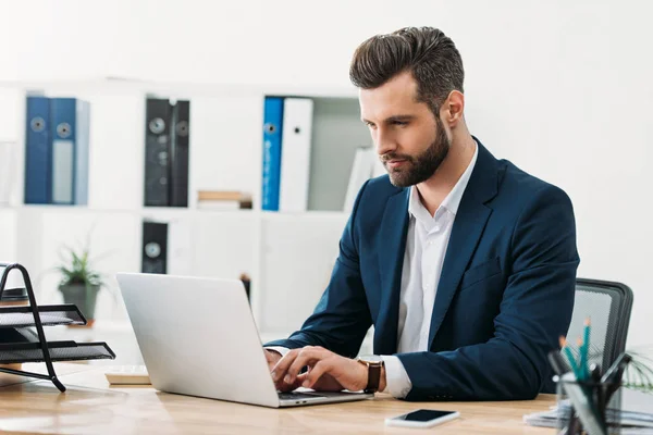 Бизнесмен в синем костюме сидит за столом с ноутбуком и печатает в офисе — стоковое фото