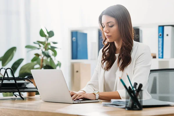 Atractiva mujer de negocios sentada en la mesa con el ordenador portátil y escribiendo en la oficina - foto de stock