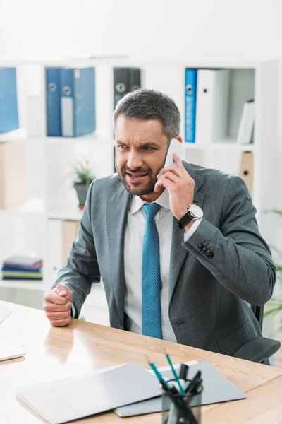 Angry businessman at table with laptop and talking on smartphone in office — Stock Photo