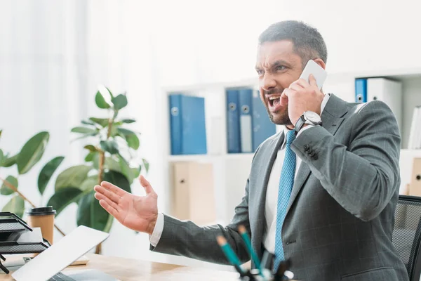 Angry businessman sitting at table with laptop and talking on smartphone in office — Stock Photo