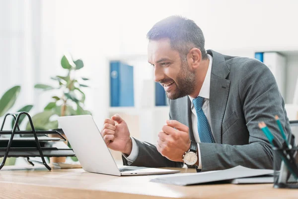 Hombre de negocios guapo sentado en la mesa, mirando a la computadora portátil y mostrando sí gesto en la oficina - foto de stock