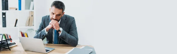 Hombre de negocios guapo en traje gris sentado en la mesa con el ordenador portátil y pensando en la oficina — Stock Photo