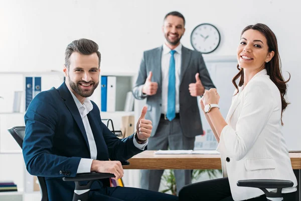 Enfoque selectivo de los inversores sonrientes y asesor guapo con pulgares en segundo plano - foto de stock