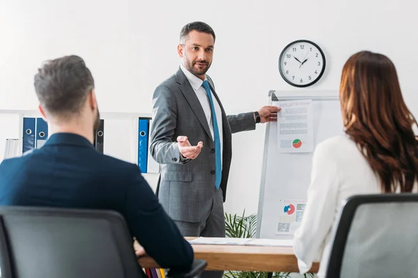 Selective focus of handsome advisor showing document to investors in suits at workplace — Stock Photo