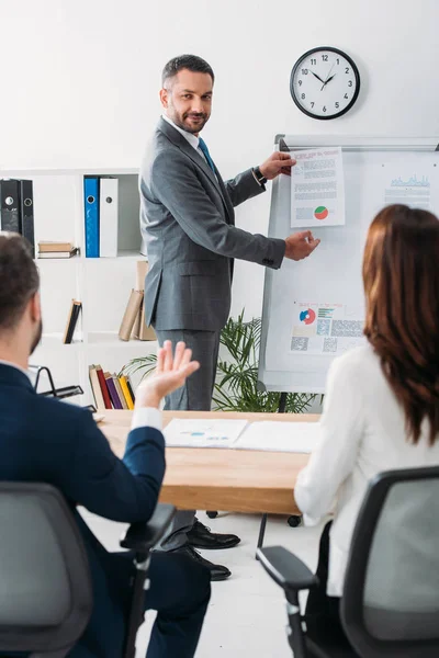 Selective focus of handsome advisor showing document to investors in suits at workspace — Stock Photo