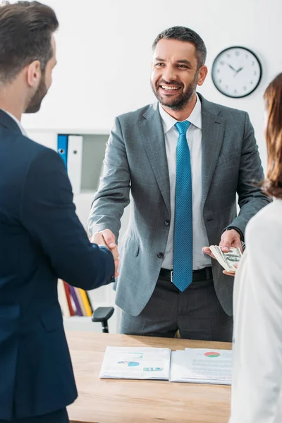 Selective focus of advisor shaking hands with investors and taking dollar banknotes at office — Stock Photo