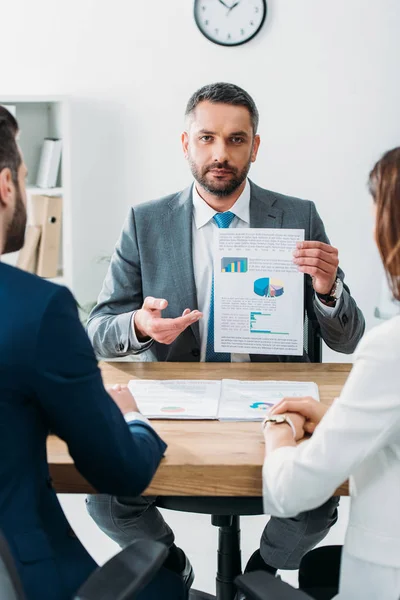 Selective focus of handsome advisor in suit showing document to investors at workspace — Stock Photo