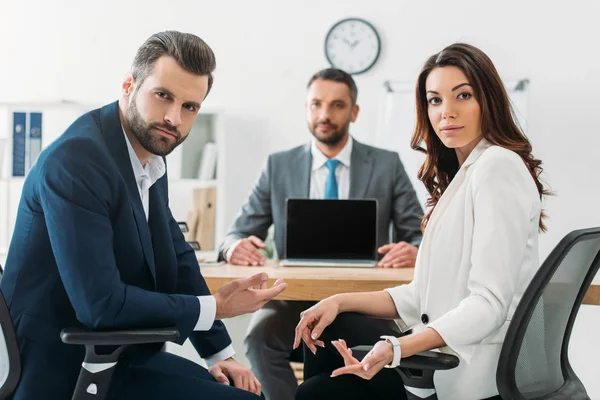 Ciblage sélectif des investisseurs en costumes regardant la caméra et le conseiller tenant un ordinateur portable avec écran blanc sur fond — Photo de stock