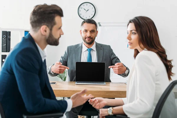 Enfoque selectivo del asesor guapo apuntando con los dedos a la computadora portátil con pantalla en blanco - foto de stock