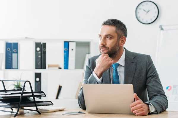 Beau conseiller en costume regardant loin avec ordinateur portable sur la table — Photo de stock