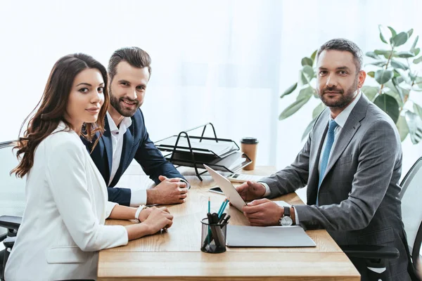 Asesor guapo en traje e inversores mirando a la cámara y sonriendo en el lugar de trabajo - foto de stock