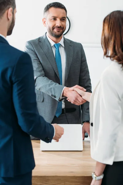 Messa a fuoco selettiva di bel consulente in giacca e cravatta e gli investitori si stringono la mano sul posto di lavoro — Foto stock