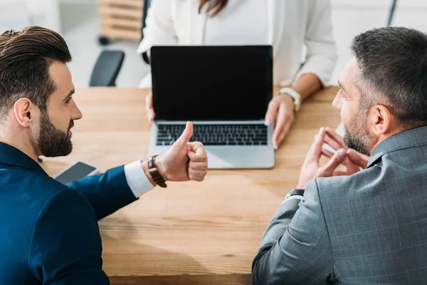 Selective focus of investors with thumb ups and advisor showing laptop with copy space at workplace — Stock Photo