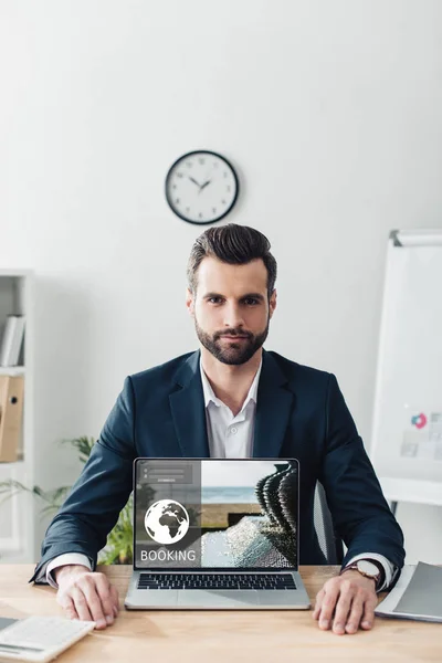Schöner Berater im Anzug zeigt Laptop mit Buchungswebseite auf dem Bildschirm — Stockfoto