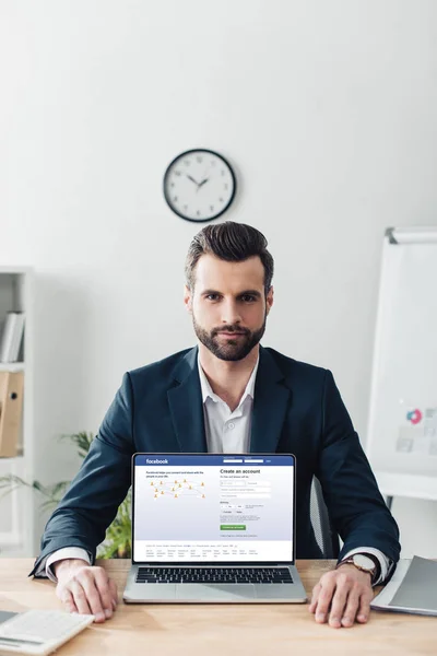 Asesor guapo en traje mostrando portátil con el sitio web de facebook en la pantalla - foto de stock