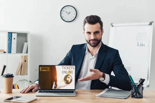 Handsome advisor in suit pointing with fingers at laptop with tickets online website on screen at office — Stock Photo