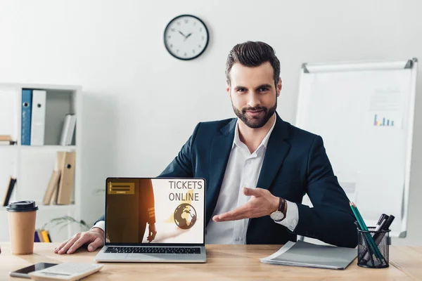 Beau conseiller en costume pointant avec les doigts à l'ordinateur portable avec des billets site web en ligne à l'écran au bureau — Photo de stock