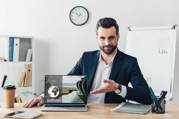 Beau conseiller en costume pointant avec les doigts à l'ordinateur portable avec site de réservation à l'écran au bureau — Photo de stock