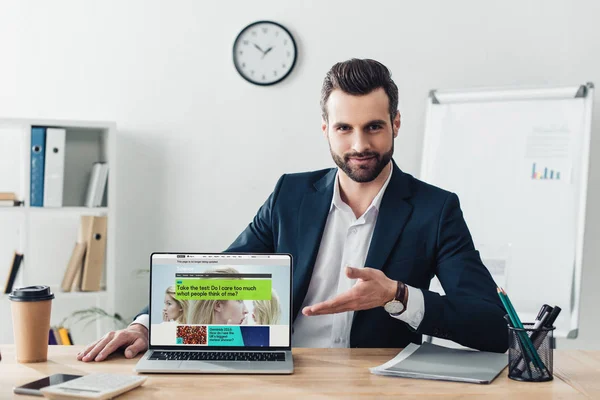 Asesor guapo en traje apuntando con los dedos a la computadora portátil con sitio web de la penes en la pantalla en la oficina - foto de stock