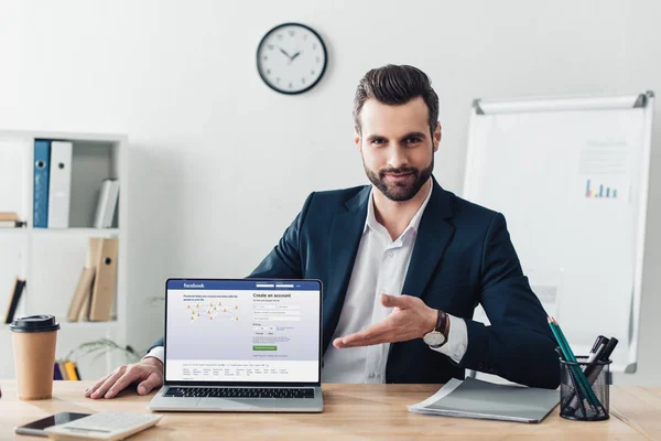 Asesor guapo en traje apuntando con los dedos a la computadora portátil con el sitio web de facebook en la pantalla en la oficina - foto de stock