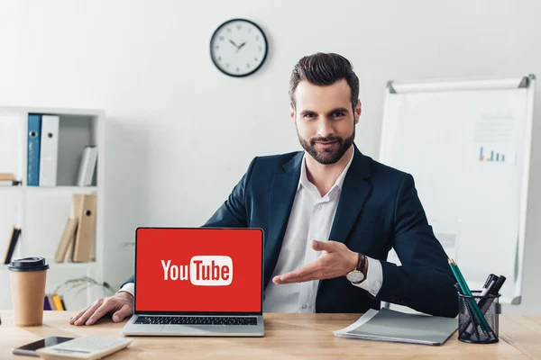 Conselheiro bonito em terno apontando com os dedos para laptop com site do youtube na tela no escritório — Fotografia de Stock