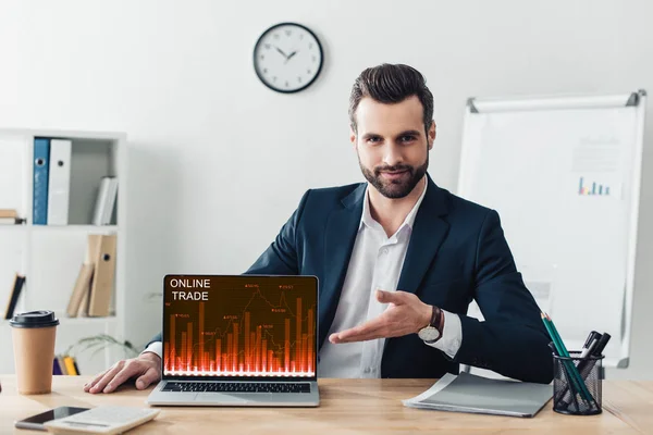 Asesor guapo en traje apuntando con los dedos a la computadora portátil con el sitio web de comercio en línea en la pantalla en la oficina - foto de stock