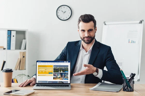 Asesor guapo en traje apuntando con los dedos a la computadora portátil con el sitio web de reserva en la pantalla en la oficina - foto de stock