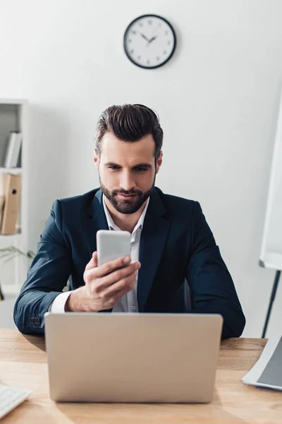 Asesor guapo en traje usando smartphone con portátil en el lugar de trabajo - foto de stock