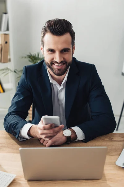 Lächelnder und gutaussehender Berater im Anzug mit Smartphone am Arbeitsplatz — Stockfoto